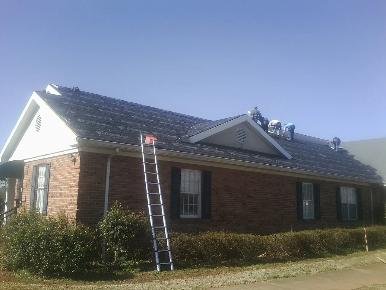 Crew tearing shingles off a brick ranch roof with a ladder set — Emergency Roof Repair work by our crew in Sumpter