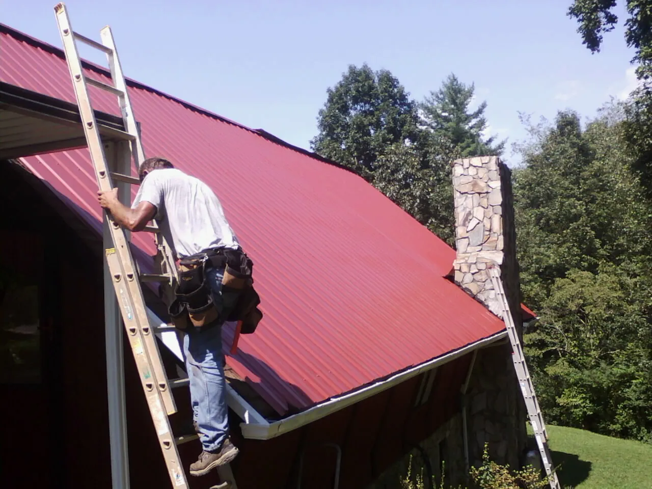 Roofer on a ladder beside a red metal roof and stone chimney — Soffit & Fascia Repair by Roofing Brothers in Sumpter